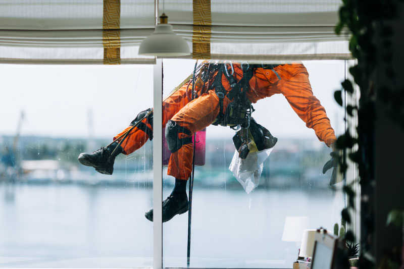 A window cleaner in orange protective gear is suspended outside a large window, using rope access construction techniques and safety equipment to clean the glass in South Africa. - OADR Training