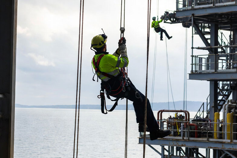 A worker using rope access hangs from ropes on the side of an offshore platform above the water, with another worker visible on the structure, showcasing skills often seen in rope access mining in South Africa. - OADR Training